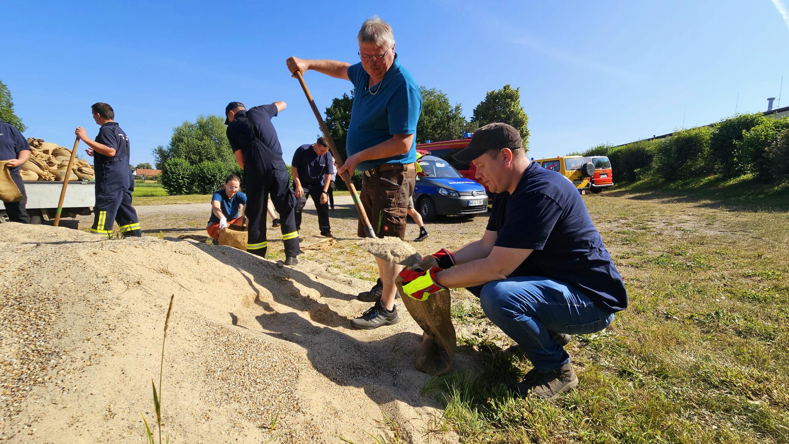 vorn links ein Berg mit Sand; dahinter die Teilnehmer der Wasserwehrschulung, welche Sandsäcke befüllen
