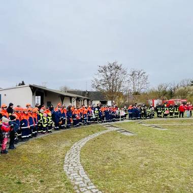 Die Feuerwehrleute und Kitakinder sind im Halbkreis aufgestellt und Hören zwei Feuerwehrleuten zu