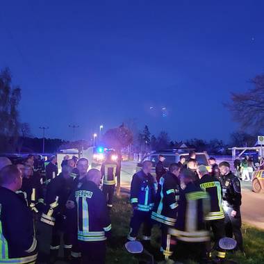links stehen Mitglieder der Feuerwehr, rechts ein Polizeiauto; der Himmel wird langsam dunkel