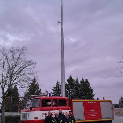 Sirene hoch auf Metallpfosten dazu rotes Feuerwehrauto und 5 Kameraden der Feuerwehr
