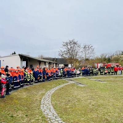 Die Feuerwehrleute und Kitakinder sind im Halbkreis aufgestellt und Hören zwei Feuerwehrleuten zu