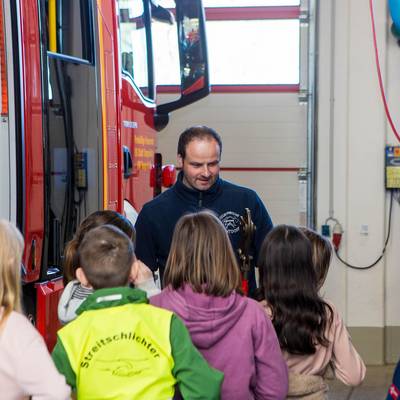 Herr Paasche zeigt den Kindern die Ausrüstung auf dem Feuerwehrauto. Links sind das Auto und Ausrüstungsteile zu erkennen.