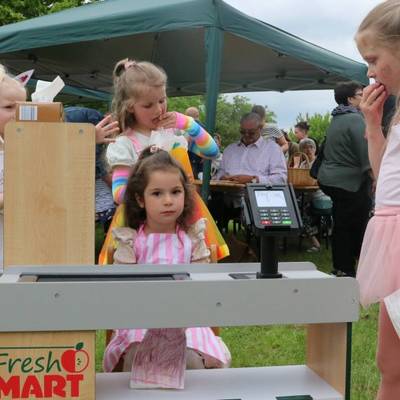 Im Vordergrund sind mehrere Kinder zu sehen, drei der Kinder stehen hinter einer Spielverkaufskasse und ein Kind steht als Interessent davor. Im Hintergrund sind Zelte und Bänke für alle Besucher zu sehen