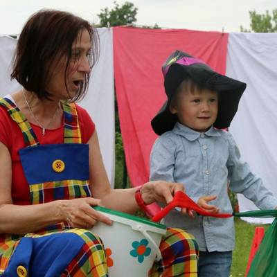 Eine Erzieherin hat sich als Clown verkleidet und zieht mit überraschtem Gesichtsausdruck ein mehrfarbiges Stofftuch aus einem Eimer. Als Assistent hilf ein Kind bei der Show