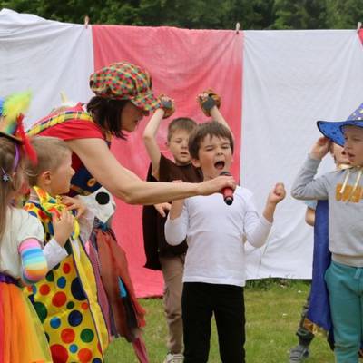 Mehrere verkleidete Kinder stehen in zwei Reihen, ein Kind in der Mitte ruf etwas in ein Mikrophon, welches von der als Clown verkleideten Erzieherin gehalten wird.