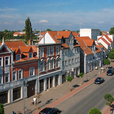 Wir sehen Tangerhüttens Bismarckstraße, es sind Fahrende und parkende Autos zu sehen, ebenso die lokalen Geschäfte.
