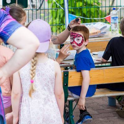 Ein Kind sitzt auf einer Bank und wird von einer KITA-Mitarbeiterin mit einem Spinnennetz im Gesicht geschminkt.